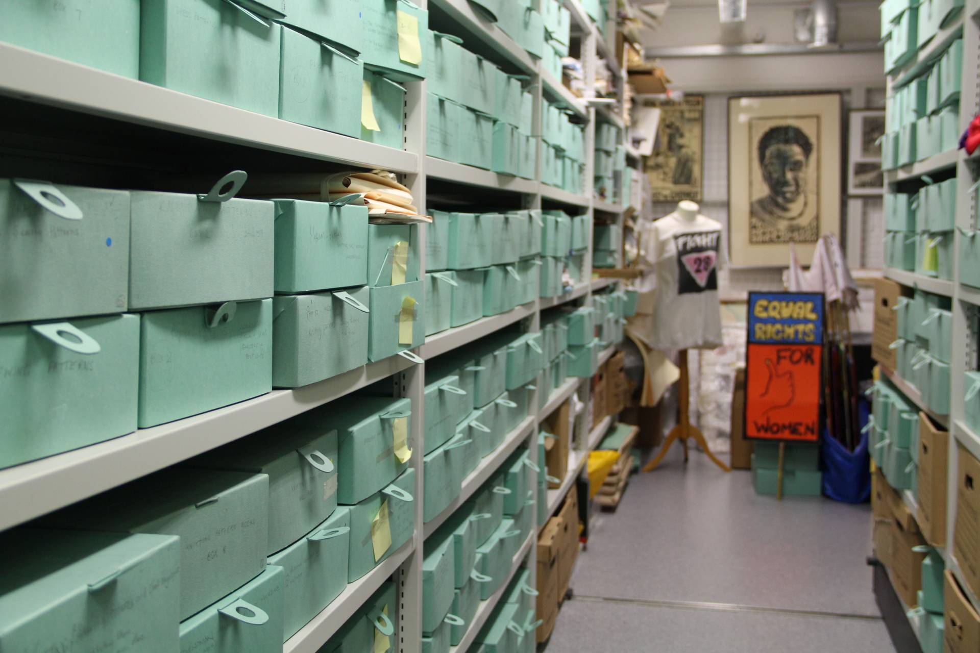 Inside one of the collections stores at GWL, with shelves full of pale green archive boxes. A T-shirt with the slogan 'Fight 28', and a placard reading 'Equal Rights for Women' are visible in the background.