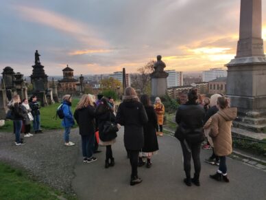 Walk attendees listen to one of our tour guides in Glasgow's Necropolis, under an evening sky