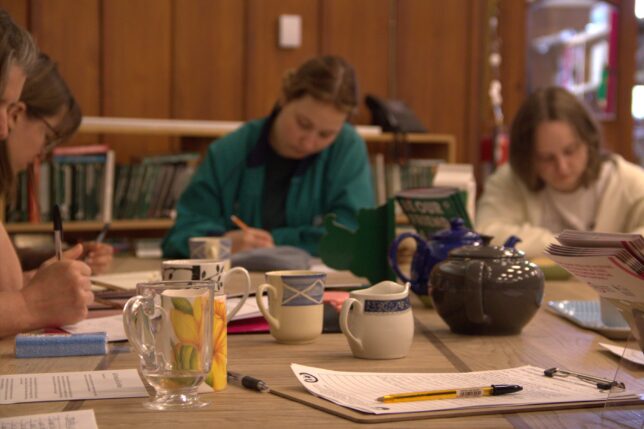 Four women sit round one end of a table, they are all looking down, writing. The table is filled with paper, mugs, teapots.