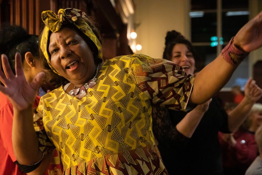 A performer in a bold yellow print dress raises her arms as she sings