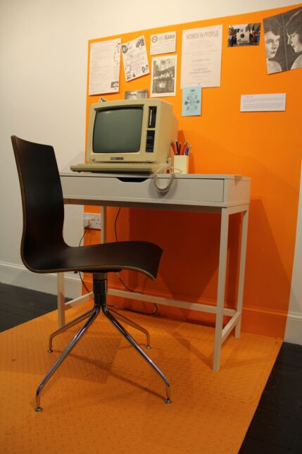 Defined orange area with desk and chair - on the desk is an Amtrad, a computer from the 1990s. A selection of black and white flyers and postcards are stuck to the wall.