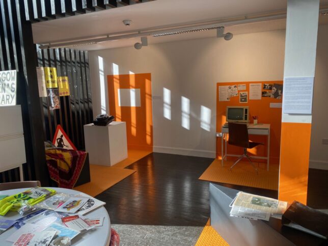 GWL Exhibition space with four distinct orange areas outlined on the floor and walls; one area has a desk with an old Amstrad PC, and another has a slide projector. GWL publicity materials are displayed throughtout the space