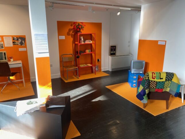 GWL Exhibition space with four distinct orange areas outlined on the floor and walls; one features a VCR combo TV and a chair, a shelf displays books and cassette tapes, an there's an old Amstrad pc on a desk