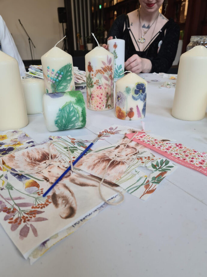 In the foreground are 7 candles, decorated with leaves and flowers, a paintbrush and a drawing of a rabbit. In the background you can see someone sat at the table with a paintbrush in their hand.