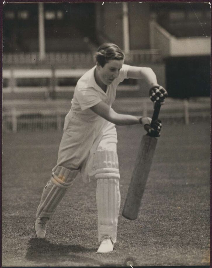 A black and white photograph of Joy Liebert. She lunges forward with her cricket bat, holding it flat in front of her.