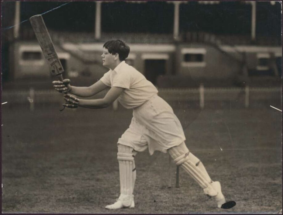 A black and white photo of Betty Archdale, her cricket bat held, mid swing in front of the cricket wicket.