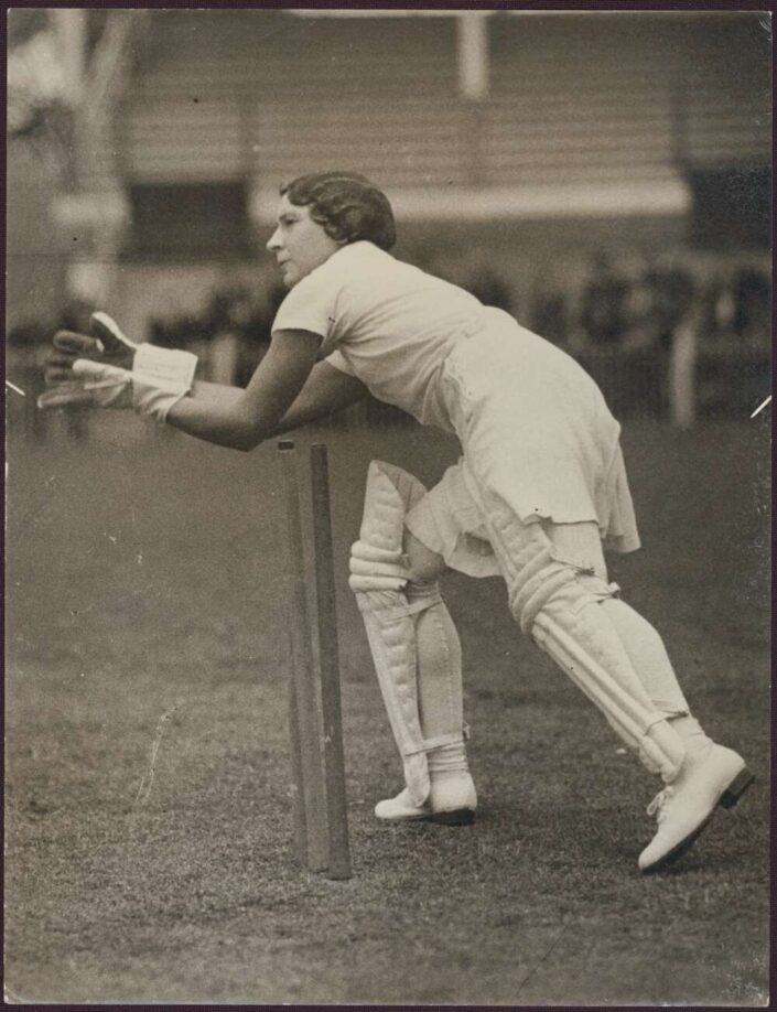 A black and white photo Betty Snowball. She stands behind the wicket and reaches with her whole body to one side, hands cupped to catch a ball.