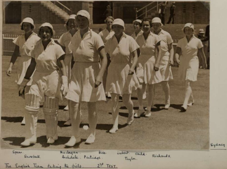 A black and white photo of the England women's cricket team from 1935 walking out onto a cricket field looking cheerful. They're all dressed in white including white stockings, white culottes and floppy white hats.