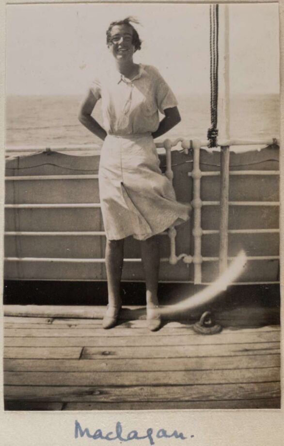 A black and white portrait photo of Myrtle Maclagan. She cheerfully poses by the railing of the ship, the sea behind her and the wind whipping her skirt and hair.