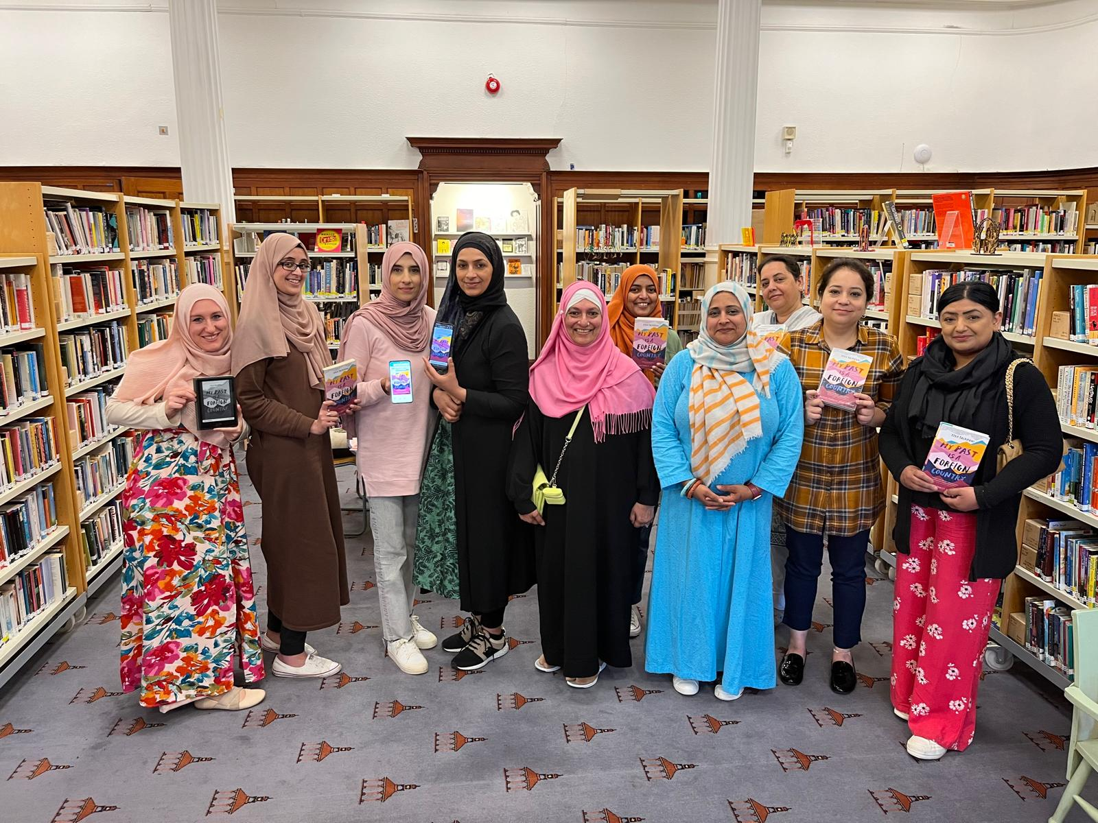 ten women stand in a line, smiling at the camera in Glasgow Women's Library. They are smiling at the camera and holding books. Some of them wear headscarves and some don't.