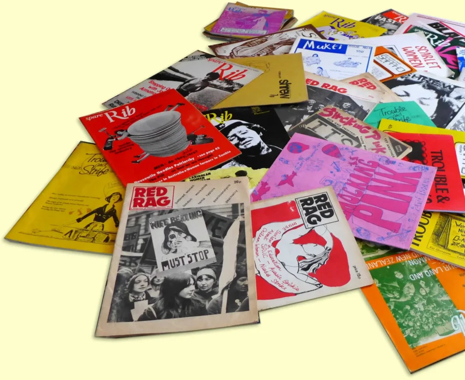 A pile of feminist magazines spilling in different directions over a plain yellow background. Titles that can be read on the covers include Red Rag, Spare Rib, Shrew, Trouble and strife, Shocking Pink, Mukti and Sappho.