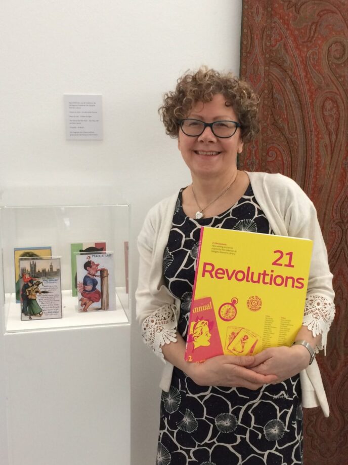 Morag Smith staning holding a copy of 21 Revolutions. She is smiling and wearing a white cardigan and short black flowery dress