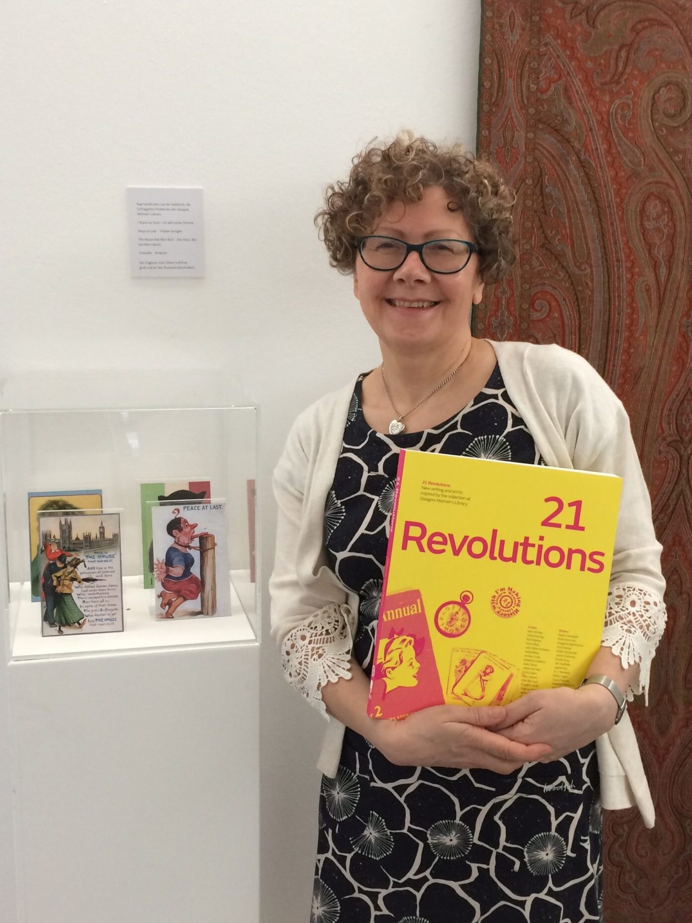 Morag Smith staning holding a copy of 21 Revolutions. She is smiling and wearing a white cardigan and short black flowery dress