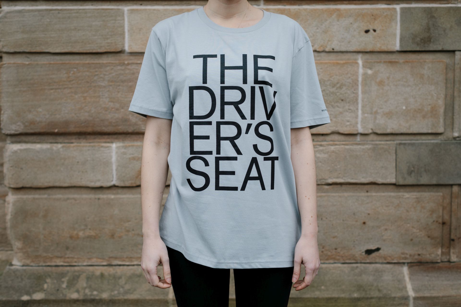 A person, visible from the neck down, wearing a grey T-shirt printed with 'The Driver's Seat' in large black letters over several lines.