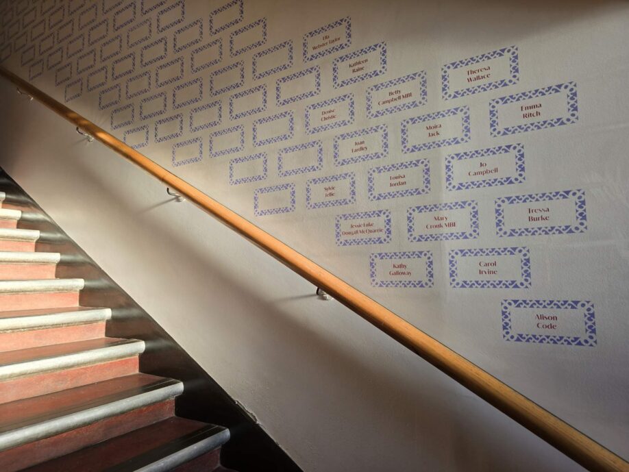 Staircase with a wooden handrail. The wall is printed with lilac blocks with names in some of them