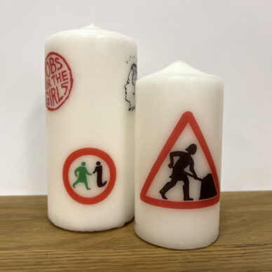 A large candle with several logos from GWL's past, next to a small candle with the 'Women at Work' logo.