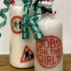 A large candle with several logos from GWL's past, next to a small candle with the 'Jobs for the Girls' logo. Both are wrapped in cellophane and tied with ribbons.