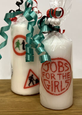 A large candle with several logos from GWL's past, next to a small candle with the 'Jobs for the Girls' logo. Both are wrapped in cellophane and tied with ribbons.