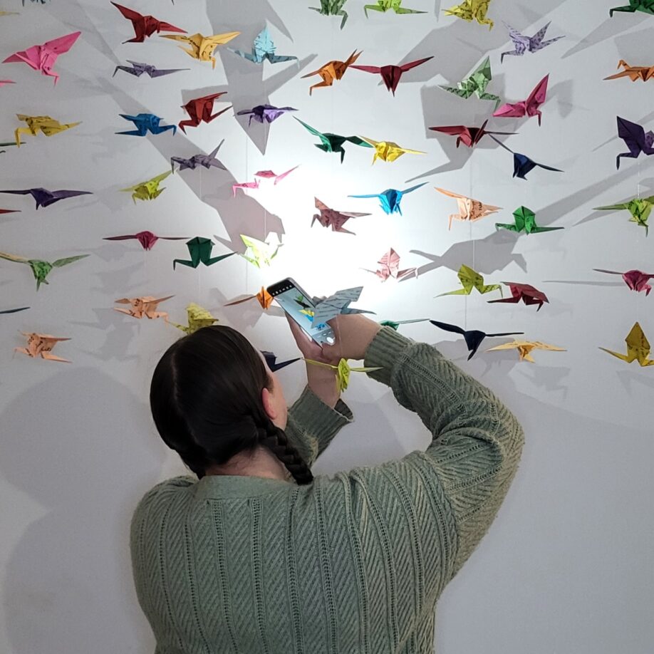 Alexandra Compton stands amongst her installation of colourful paper cranes, with her back to the camera, the flash from her phone camera casting shadows against a white wall.