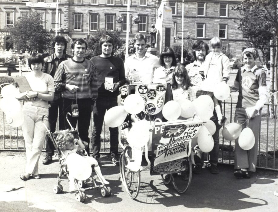 Black and white photograph of a group of 11 people made up of adults and children all gathered for the photo. They have an old fashioned pram covered in badges and a banner reading ‘War is not healthy for children and other living things’. Their hairstyles and clothes indicate it is the late 70s/early 80s.