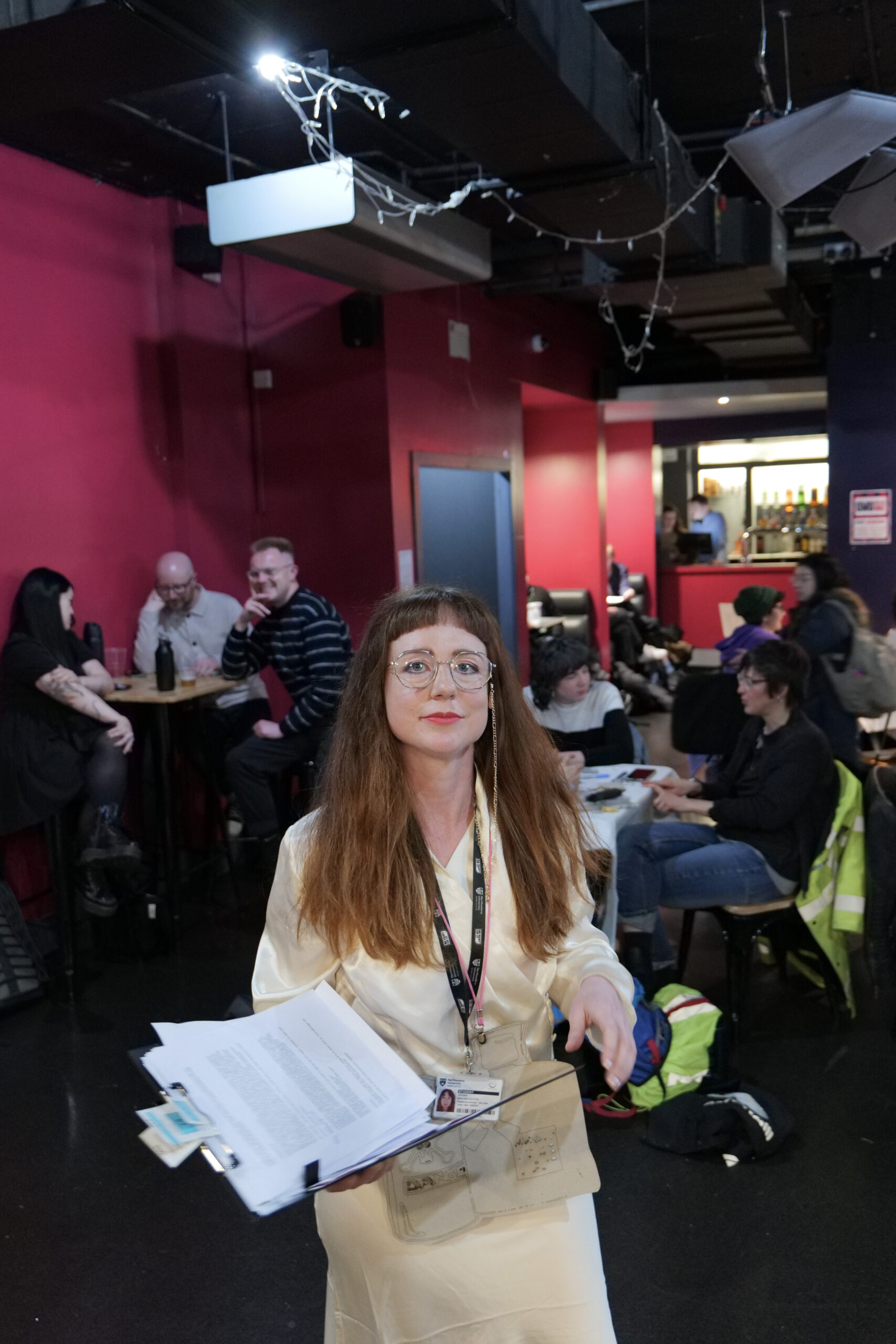 Shona Macnaughton in the performance Give Your Thoughts and Win, at the University of Glasgow Shona McNaughton stands, facing the camera holding a clipboard with documents, wearing a lanyard and glasses with a chain attached. She’s wearing a white dress. In the background groups of people are sitting at tables in casual dress.