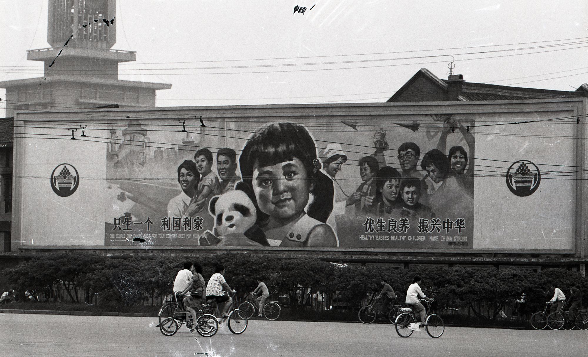 Black and white photograph of a billboard with a a girl at the forefront holding a panda. the billboard has chinese writing on it , which translated into english reads, 'Healthy babies, Healthy Children make China Strong. In front of the billboard is 7 people on bicycles riding in different directions.