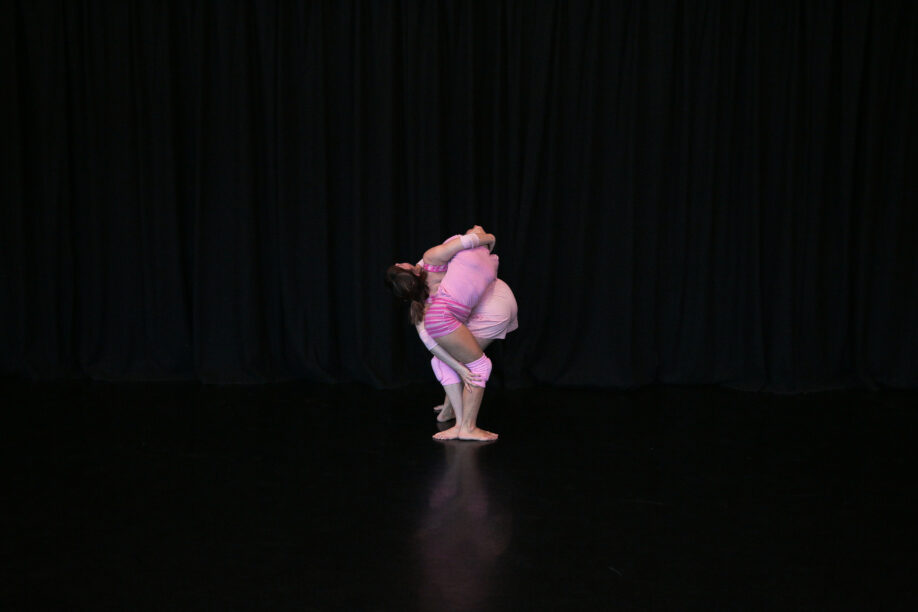 Two bodies are holding each other close and supporting each other by balancing their weight. They are both wearing pink and have twisted their forms together in front of a black floor length curtain and standing on a black floor.