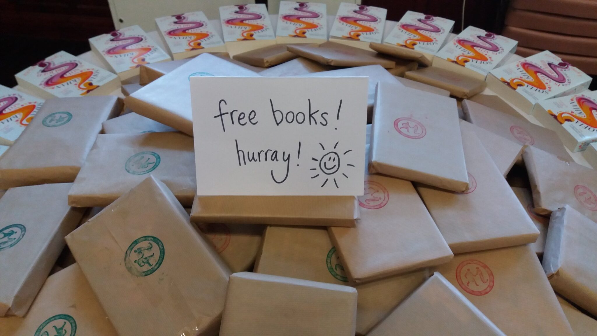 Books wrapped in brown paper with the GWL logo stamp are piled on a table. A sign on top of the pile reads 'Free books hurray!'