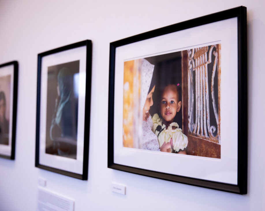 A white wall with framed photographs hanging on it. Photograph in the foreground shows a woman in a headscarf holding a young child and smiling at them. The child looks at the camera, with a slight smile. They are in a doorway, the door is half open.