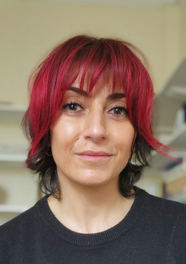 A portrait of Ebtihal Mahadeen, a feminist academic. She has short black hair, with a red fringe, and wears a nose ring.