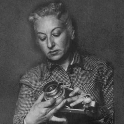 A black and white photograph of Mexican photographer, Lola Alvarez Bravo, in which she holds a camera and is looking intently at it.