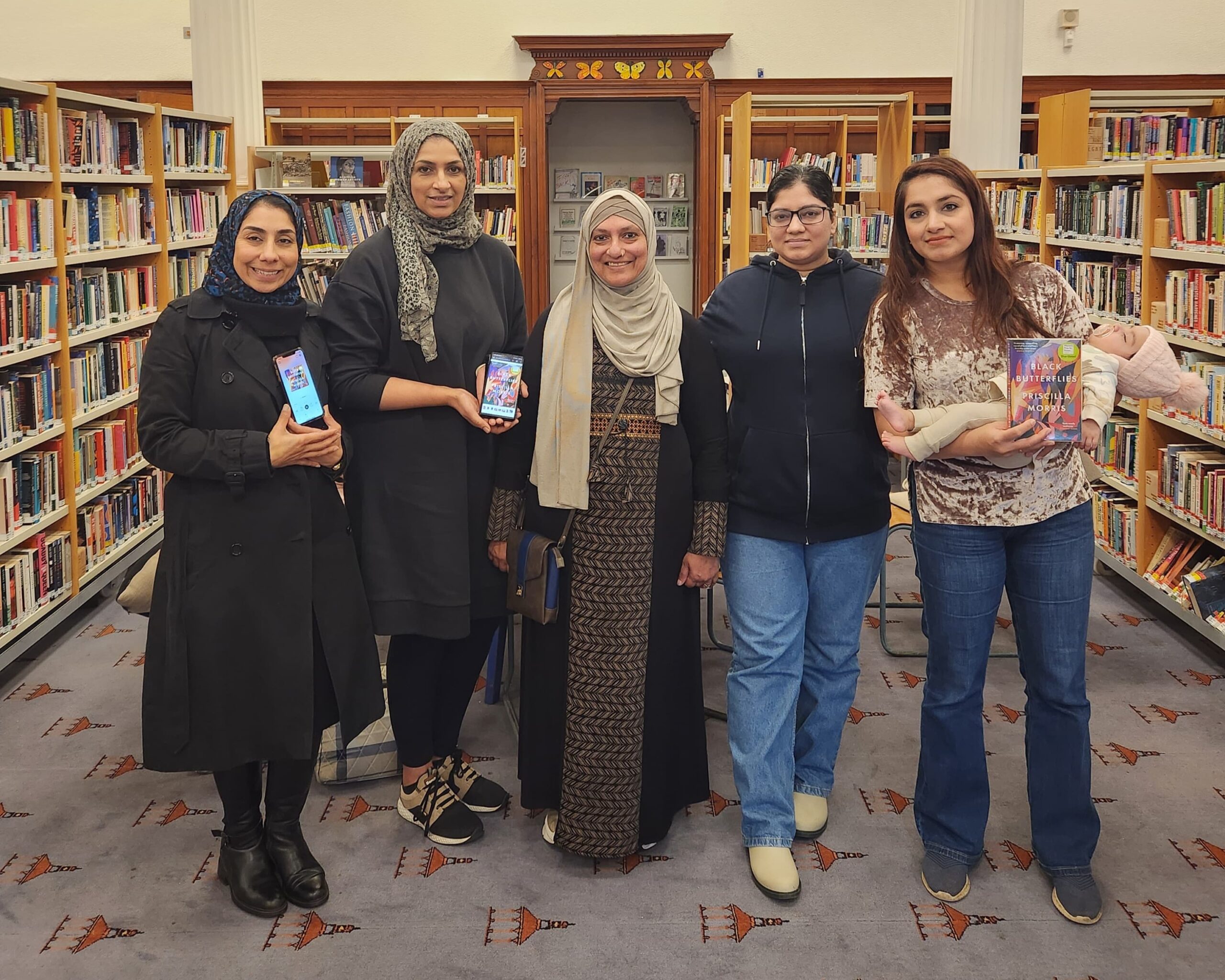 Members of GWL's Reading Group for Muslim Women stand in a line in the GWL library space, showing the camera a copy of Black Butterflies by Priscilla Morris, in physical form or on their phone screens.