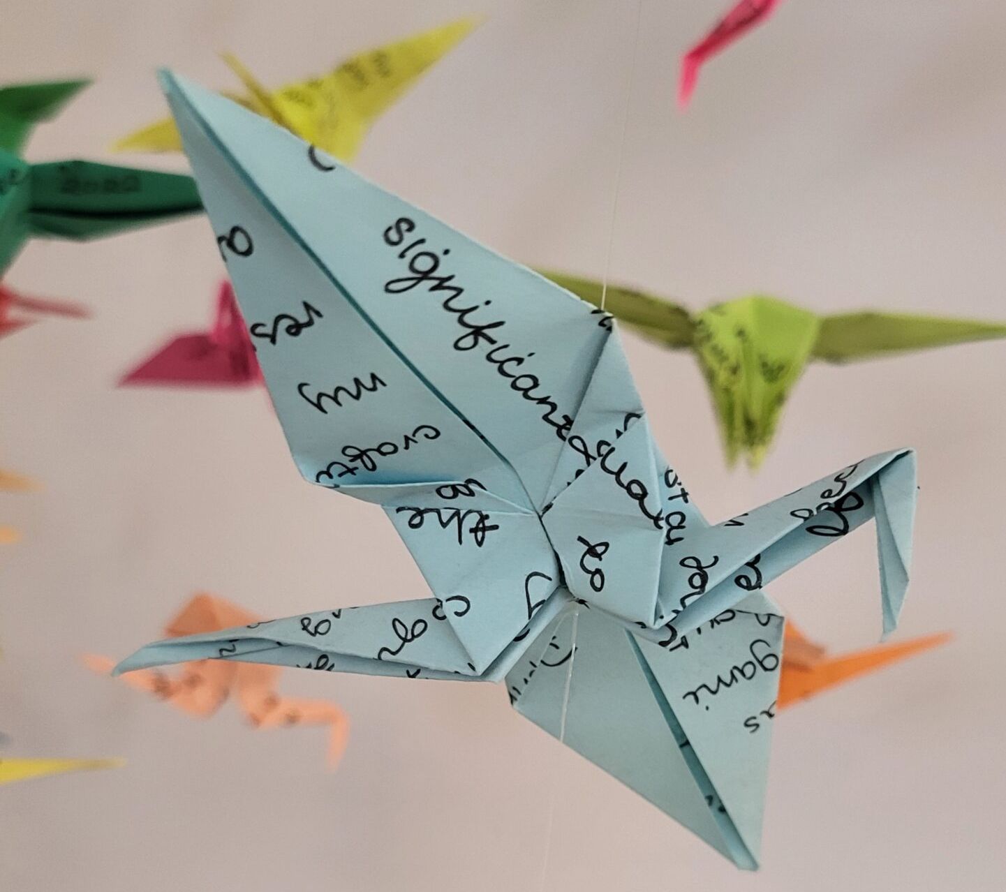 An origami crane made from blue paper covered in handwriting, hung on a thread. More cranes of different colours hang in the background.