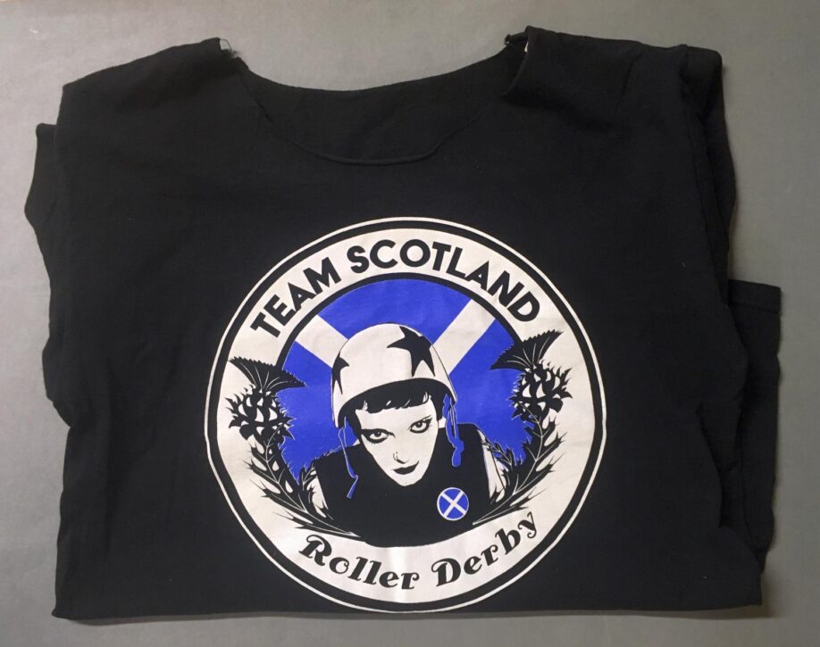 Black t-shirt with cut-away neck with a circular motif with "Team Scotland Roller Derby" around the edges and a female roller derby over a Scotland flag in the middle with thistle on either side of her