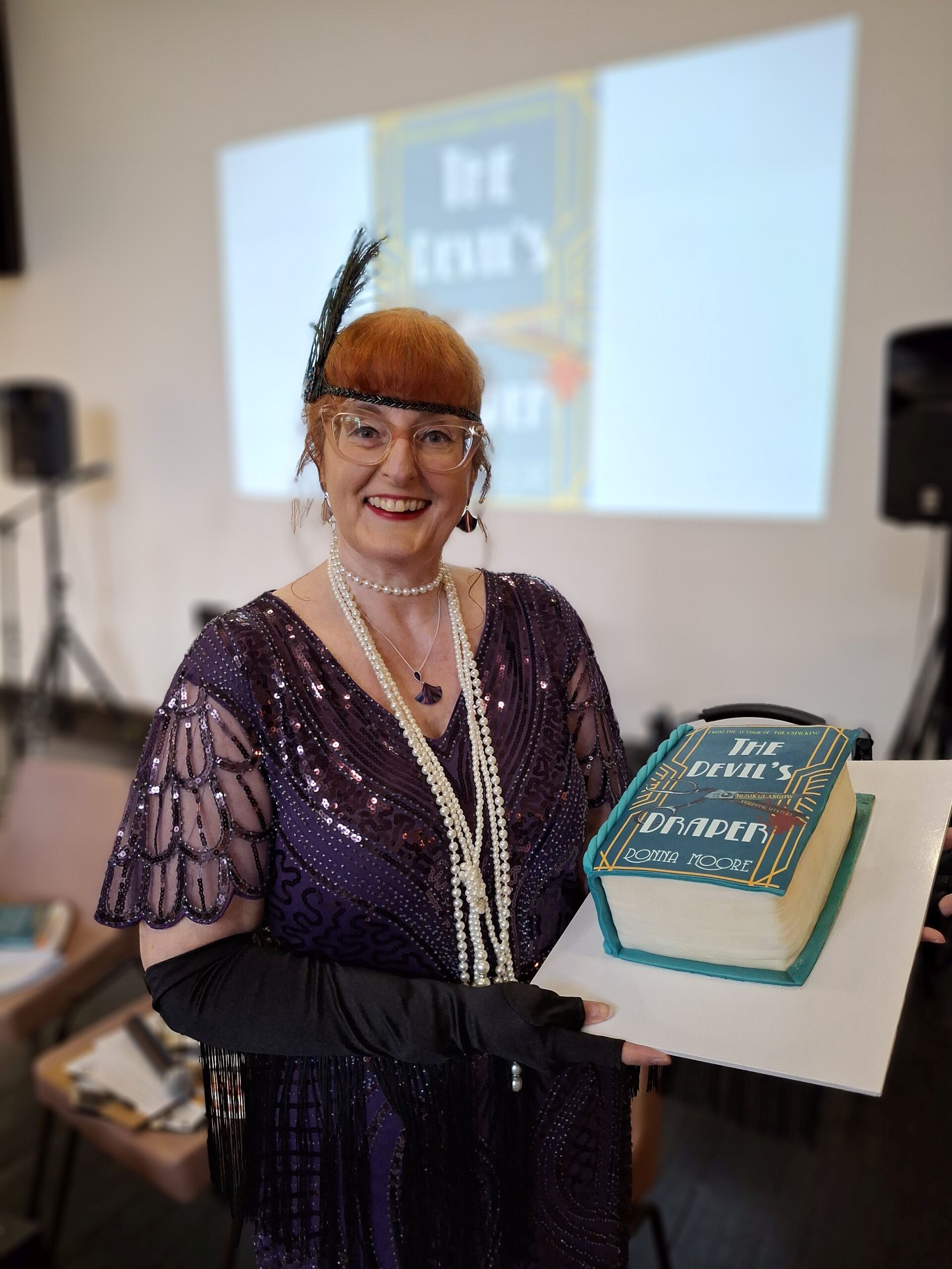 Writer Donna Moore, dressed in a 1920s flapper outfit, holds a book-shaped cake, with the book cover of A Devil's Draper on it.