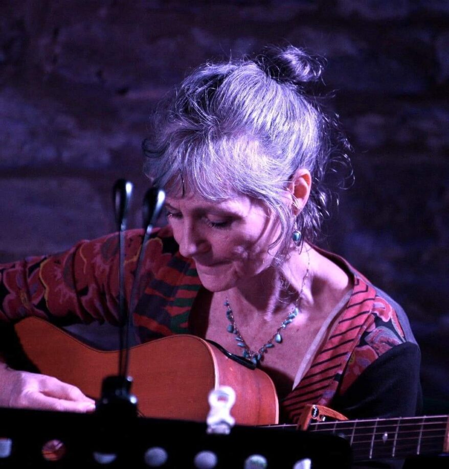 Lesley O'Brian plays guitar and sits in front of a mic stand, she is looking down at her fingers playing guitar and has her long grey hair tied up in a knot